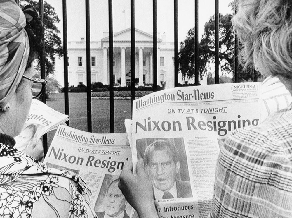 Two people outside the White House fence holding newspapers with the headline 'Nixon Resigning'